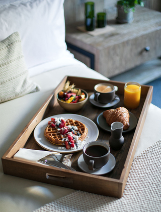 1 hotel sf waffle, croissant, fruit bowl, and various drinks on a wooden breakfast tray sitting on the guestroom bed