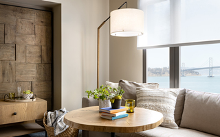 1 hotel sf guestroom living room featuring the beige sofa, solid oak top table with exposed grain, and arched floor lamp by the window with a view of the bay bridge