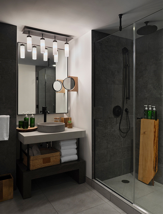 1 hotel sf concrete counter top with distressed oak vanity base. To the right is a shower with glass doors and a wooden shower caddy inside