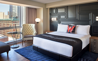 w boston guestroom bed with wingback armchair, round glass table, and a window view of the city