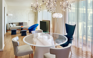 the mondrian glass round dining table in front of the living room sofa, blue wingback chair, and ottomans alongside the tv stand