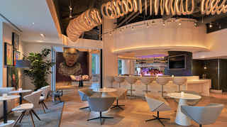 hotel zena fig leaf bar surrounded by table seating and up above hangs a wooden light installation of a serpent. On the walls, portraits of women by various female artists are displayed along with a large installation of Ruth Bader Ginsberg by artist Julie Coyle