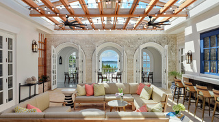 rosewood bermuda conservatory bar and lounge featuring two L shaped sofas at center, bar seating to the right, and in the back is three arched doors leading to more seating by the juliet balconies