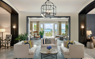 rosewood bermuda lobby featuring a glass chandelier hanging from the ceiling above 3 beige sofas and a wood coffee table at center. In the back is an adjoined room with additional seating and a balcony for guests to enjoy the view of the ocean