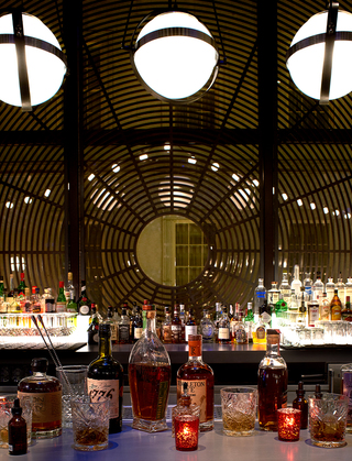 dirty habit dc liquor bottles and glasses are aligned on the bar tables. In the back, 3 circular bulbs hang from the ceiling and a black metal panel shaped as a spider web covers the window