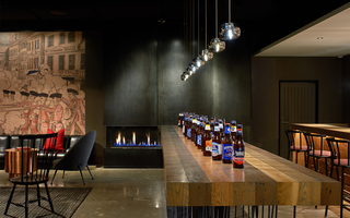 revere hotel indoor electric fireplace against the wall, in front sits a long wooden table with beer bottles lined up parallel to the bar, and to the left of the fireplace is a mural on the wall of a revolutionary war battle scene