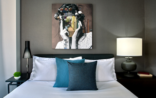 hotel zelos guestroom bed and on the wall is a mural of a woman with a numerous amount of items decorated in her hair including a cctv camera pointing from her hair to her face which is covered by the view of a city sidewalk