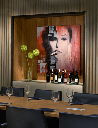 hotel palomar lounge featuring a mural of a woman with a cigarette in her mouth hanging above a cabinet with wine glasses on it. In front of the cabinet, is a long wooden rectangular dining table and black armchairs running along both sides of the table