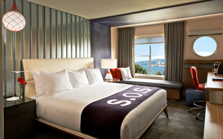 hotel zephyr guestroom bed featuring a bed cover saying S.O.S, seating by the window with an ocean view, and a wood desk with ample space