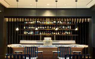 the logan lobby bar featuring 3 tiers of floating shelves filled with rows of liquor bottles and a wood carved bar table with a lit-up tabletop