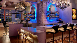 lizzie starlight pointed arch top rectangular mirrors with neon blue lights radiating light on the back of them hanging on each of the adjacent walls in the bar. On each side of the bar there are also floating shelves in the middle of both mirrors and hanging from the ceiling two metal chandeliers light up the room