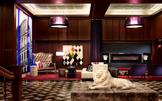 the grand hotel lobby featuring at front, a lion sculpture laying in the middle of an ottoman, and to the back wall is a long electric fireplace with a black stone mantel and a mural of the colosseum