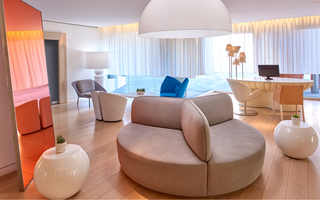 the mondrian full-length orange iridescent mirror up against the wall in front of a round oversized grey lounge chair in the lobby. Hanging above the oversized lounge chair is a large white dome pendant light. In the foreground is a concierge desk with feather light fixtures installed on the counter