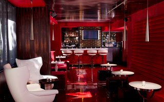 Bar rouge featuring a couple of white armchairs and tables at front and white bar stools with a small bar and a tv in the back surrounded by red floors, walls, and furniture