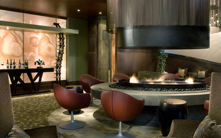 Hotel 1000 large indoor fire pit with a metal cylindrical hood and a metal mesh curtain surrounding the hood. Around the perimeter of the firepit are armchairs and additional seating in the lounge